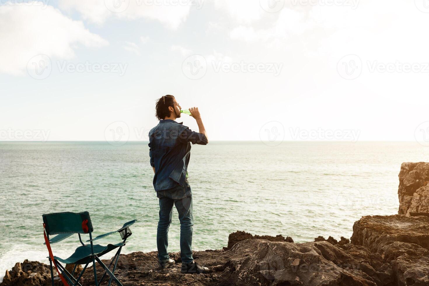 A man stands on a rocky cliff, facing the ocean and sipping a drink, as the sun begins to set. A nearby folding chair adds to the serene atmosphere, creating a sense of calmness and leisure. photo