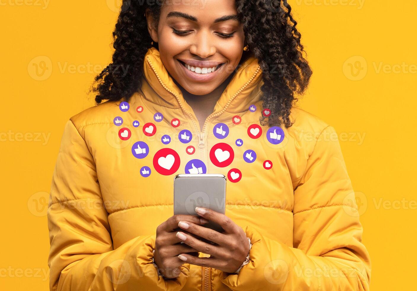 Happy Black Woman Holding Smartphone With Hearts And Like Buttons Standing On Yellow Studio Background. Phone User Networking Online Using Social Media, Reading Feed News, Commenting And Sharing Posts photo
