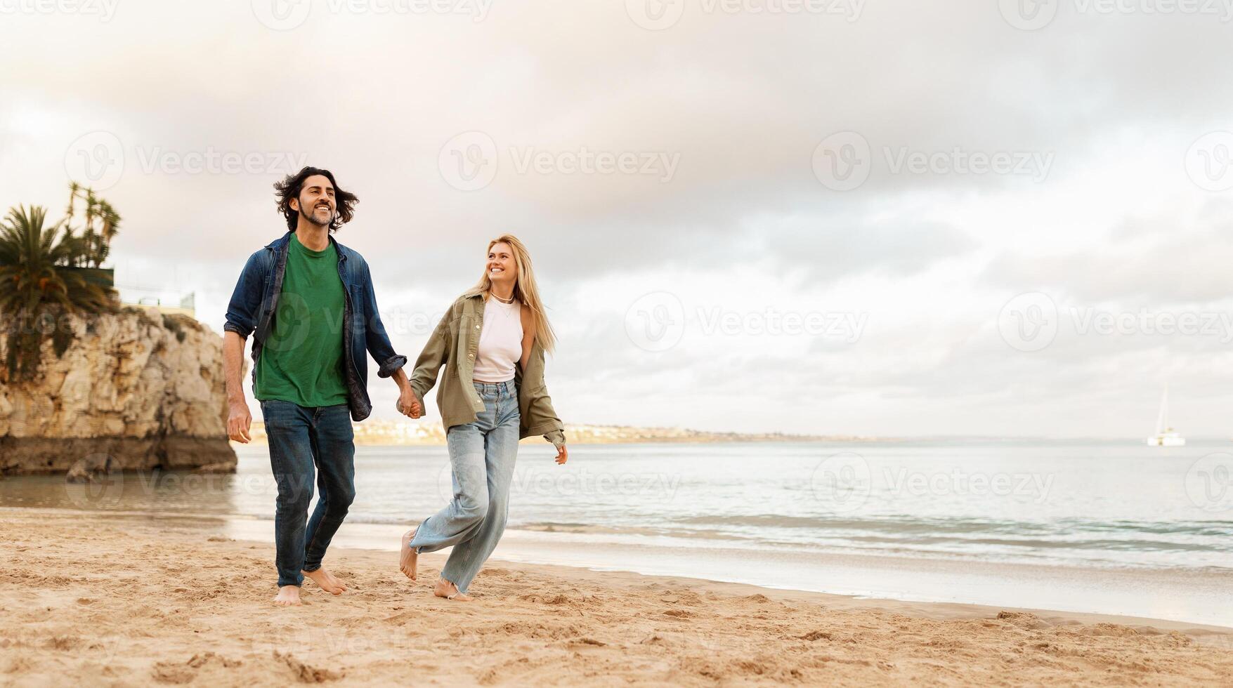 A man and woman stroll barefoot on the sandy beach, holding hands as they enjoy each other's company. The sun sets behind the clouds, casting a warm glow on the peaceful shoreline, copy space photo