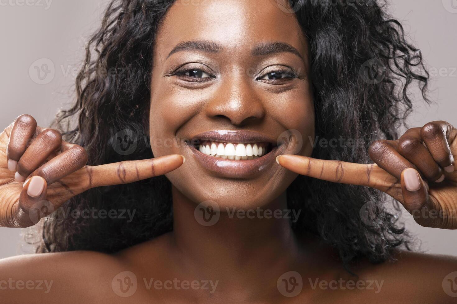 Teeth alignment and whitening concept. Happy african american girl pointing at her wide smile with two forefingers over grey background, closeup photo