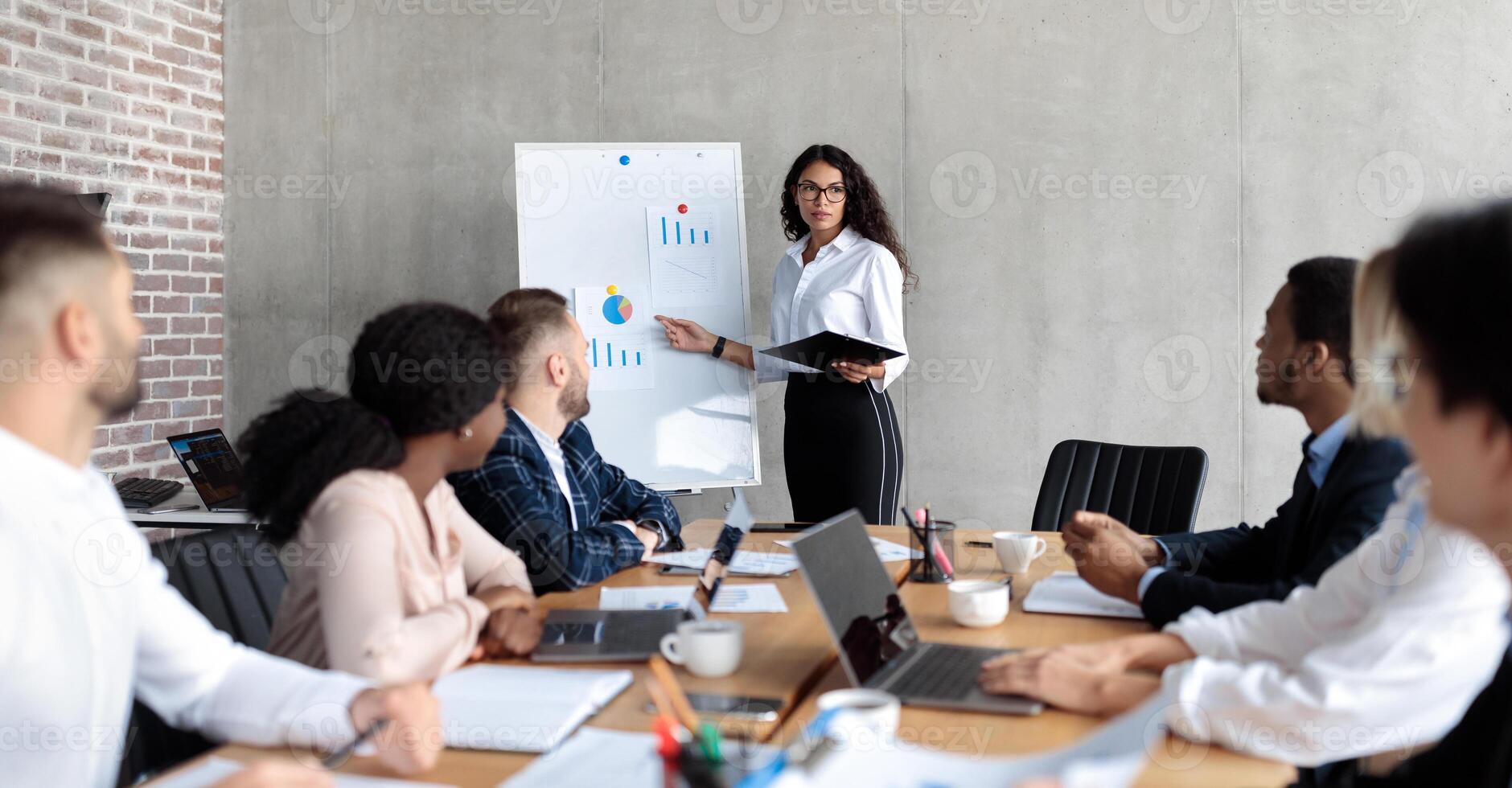 Latin Businesswoman Making Business Presentation Giving Speech For Colleagues Standing And Pointing At Blackboard With Charts And Graphs In Modern Office. Panorama, Selective Focus photo
