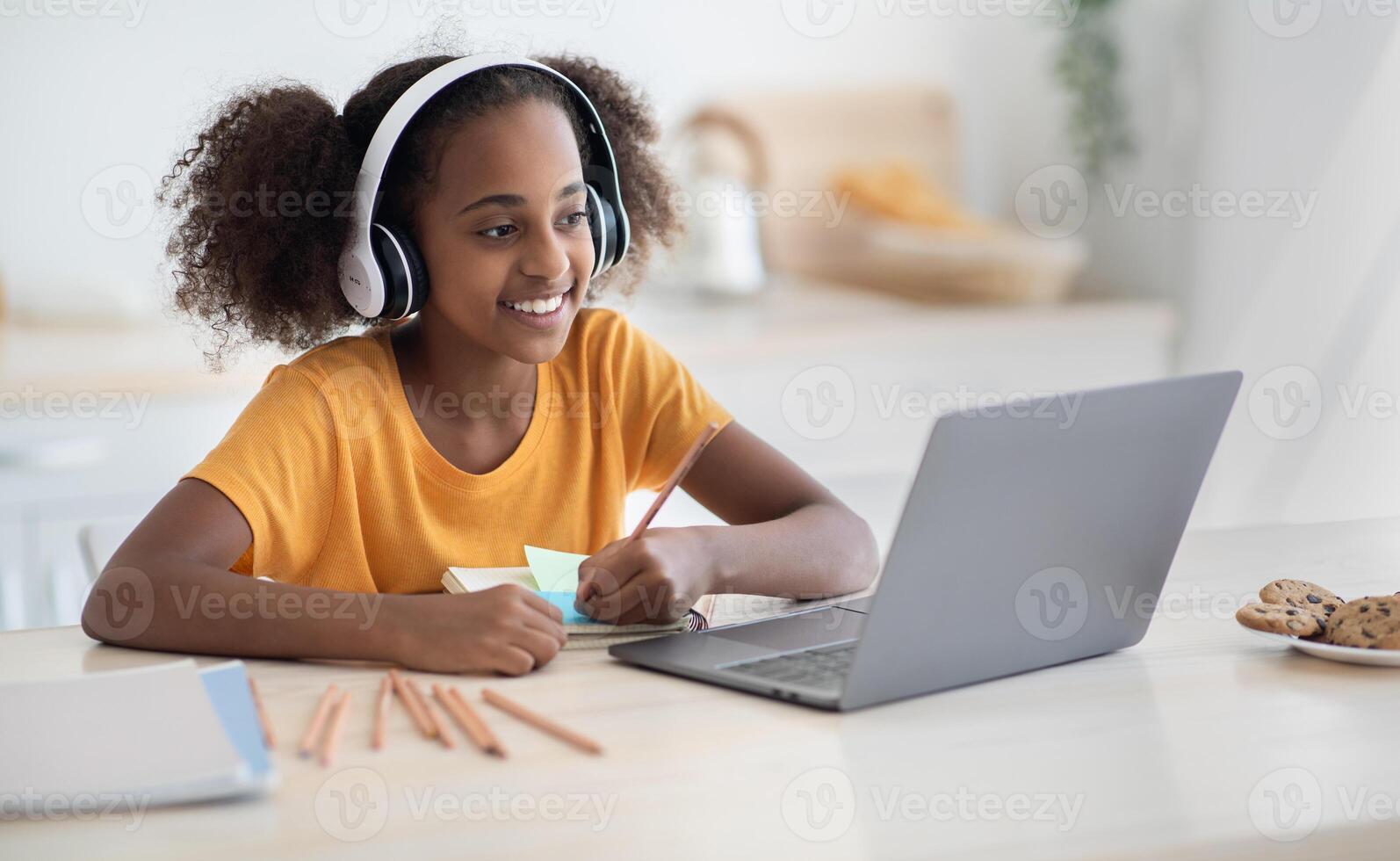 Cheerful black girl studying online, using laptop and headset at home, copy space. Cute african american teen kid having online lesson, drawing, kitchen interior. Home schooling concept photo