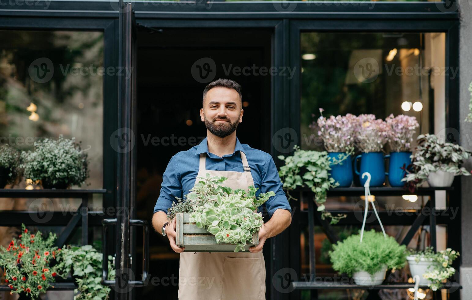 Small business owner works in flower shop and customer service. Confident smiling young bearded european male in apron carries wooden box with plants, on shop or eco cafe background, empty space photo