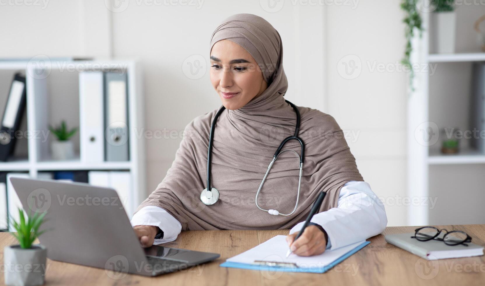 Young woman doctor having online appointment, sitting in office, using laptop and taking notes, copy space. Attractive muslim lady physician working with computer, clinic interior, remote healthcare photo