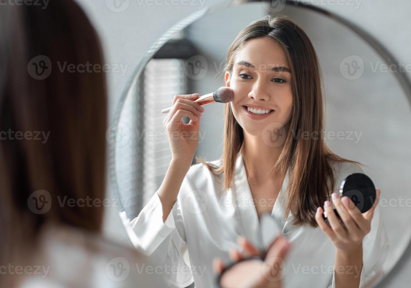 Attractive Young Woman Doing Daily Makeup While Standing Near Mirror In Bathroom, Happy Beautiful Female Putting Blush On Cheeks While Getting Ready At Home, Selective Focus On Reflection photo