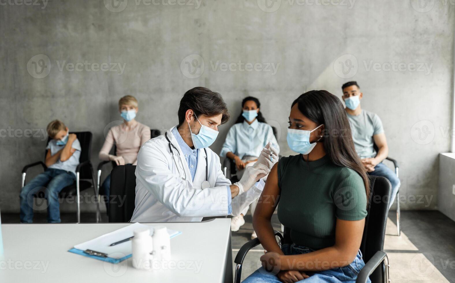 African american young woman getting vaccinated against COVID-19 at vaccination walk-in site, international group of patients waiting for immunization while coronavirus pandemic at hospital photo
