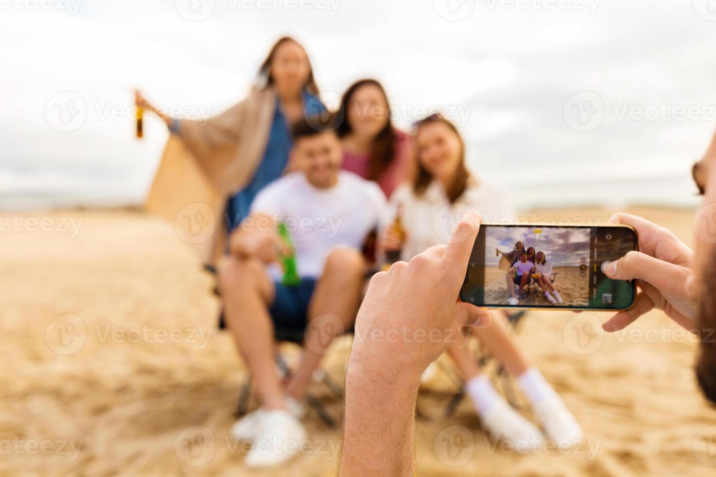 A group of friends poses joyfully on the beach, holding drinks and smiling as one of them takes a picture with a smartphone during sunset. The atmosphere is filled with laughter and happiness. photo