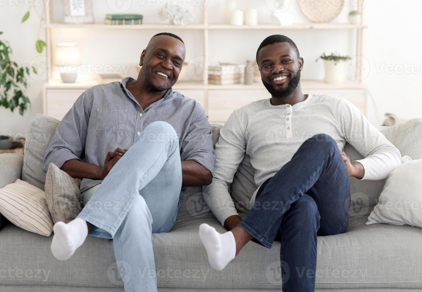 Like Father Like Son. Happy Black Millennial Man Sitting In Similar Pose On Couch With His Dad, Smiling At Camera, Relaxing At Home Together photo