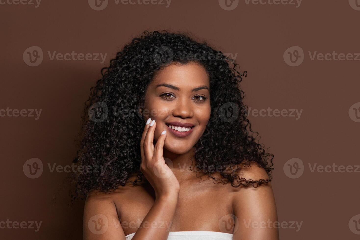 A young woman with curly hair is smiling confidently while resting her hand on her face. She is dressed in a simple top, creating a calming atmosphere with a rich brown backdrop. photo