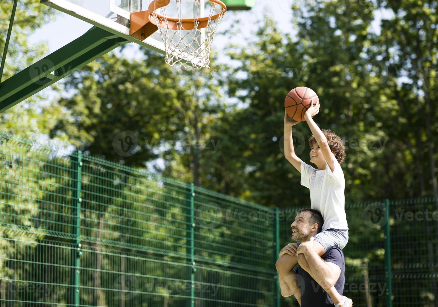 Happy father holding his little son on shoulders, helping him to score a basket on a playground photo