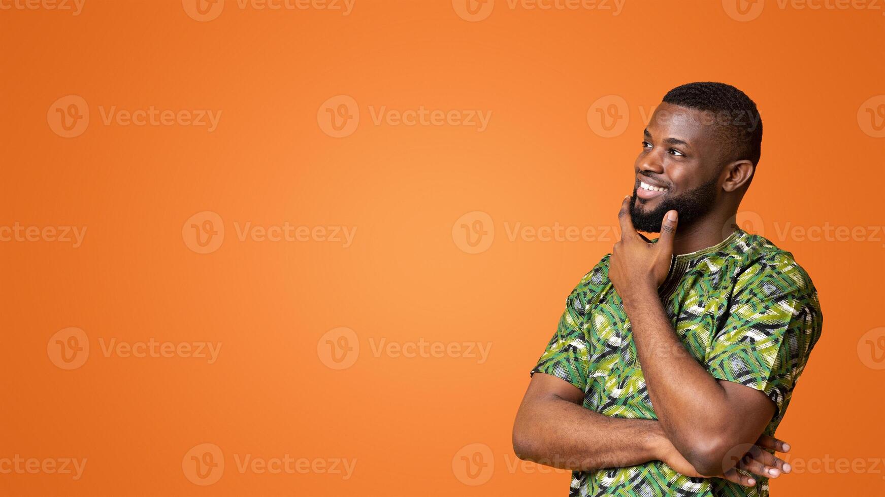 Curious black guy in african green costume touching his chin, looking at copy space, interested in nice proposition, yellow sudio background, panorama photo