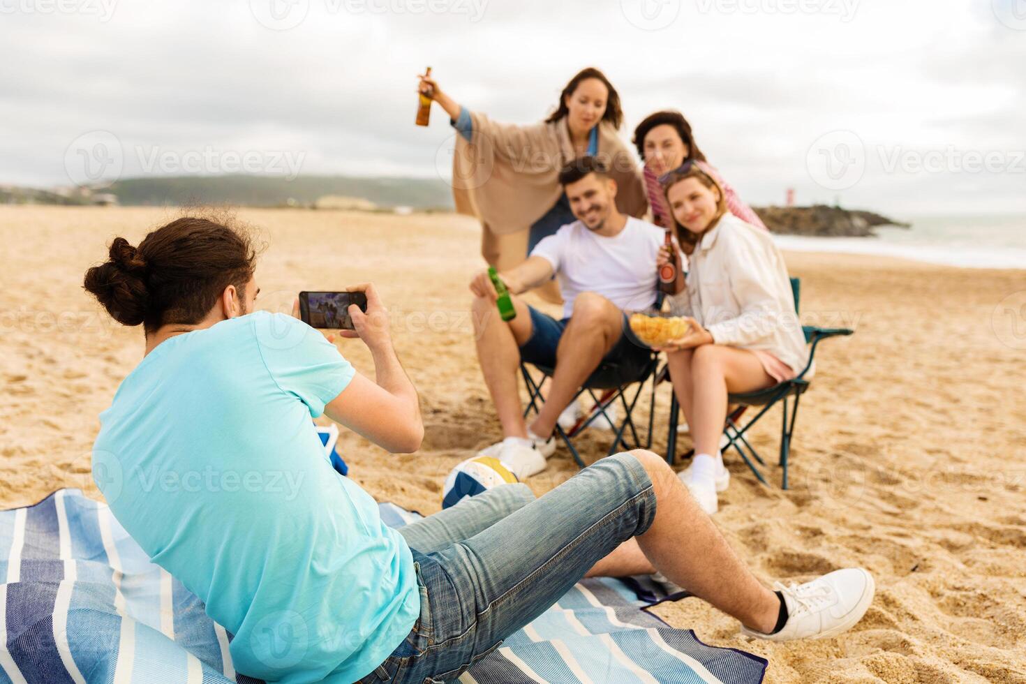 A group of five friends relaxes on the beach while enjoying snacks and drinks. One friend captures the moment on his phone, creating lasting memories of their summer outing. photo