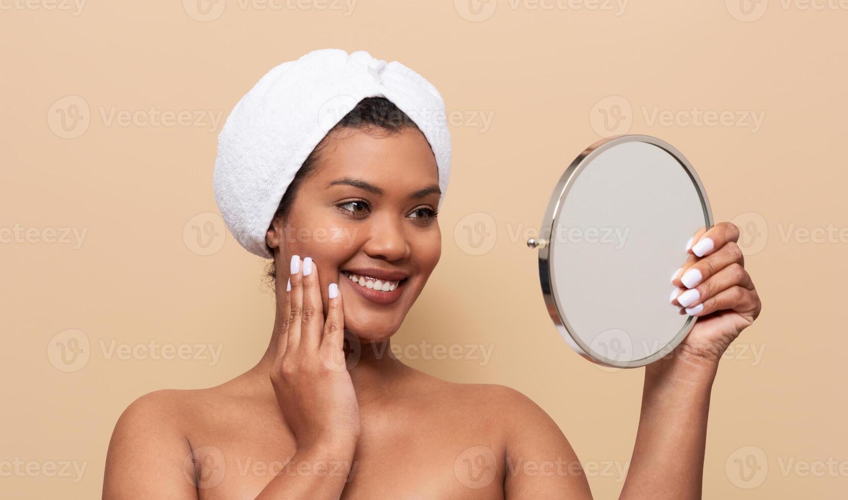 A young woman with a towel on her head smiles while looking at her reflection in a small round mirror. She touches her face, showcasing her glowing skin after a skincare routine. photo