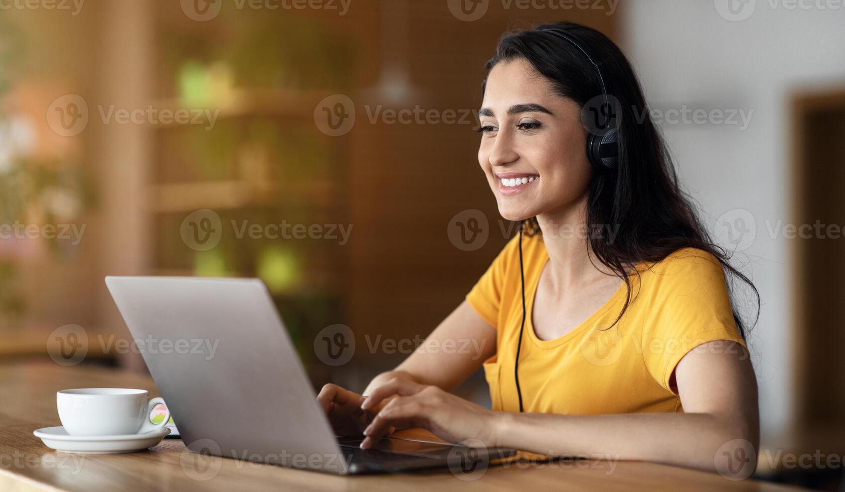 Joyful girl student studying online at cafe, using laptop and earphones, empty space photo