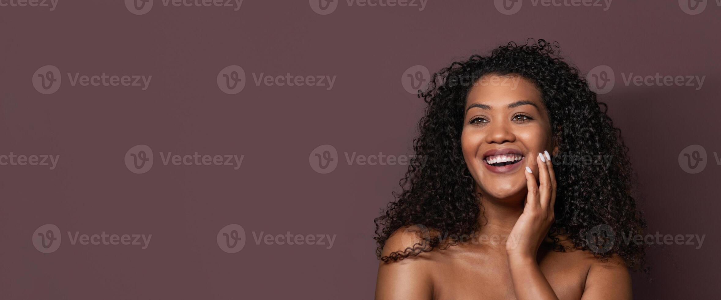 A joyful young woman with curly hair smiles confidently while resting her hand on her face. The solid purple background adds warmth to her cheerful expression, perfect for uplifting visuals. photo