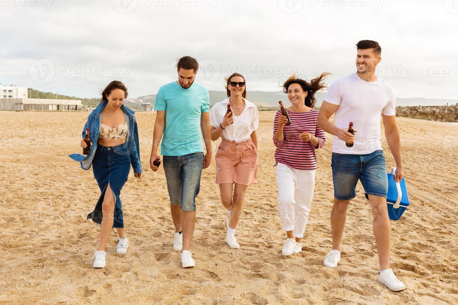 A group of five friends strolls along the sandy beach, chatting and holding drinks. The sun shines brightly as they enjoy their time together, creating a joyful atmosphere. photo