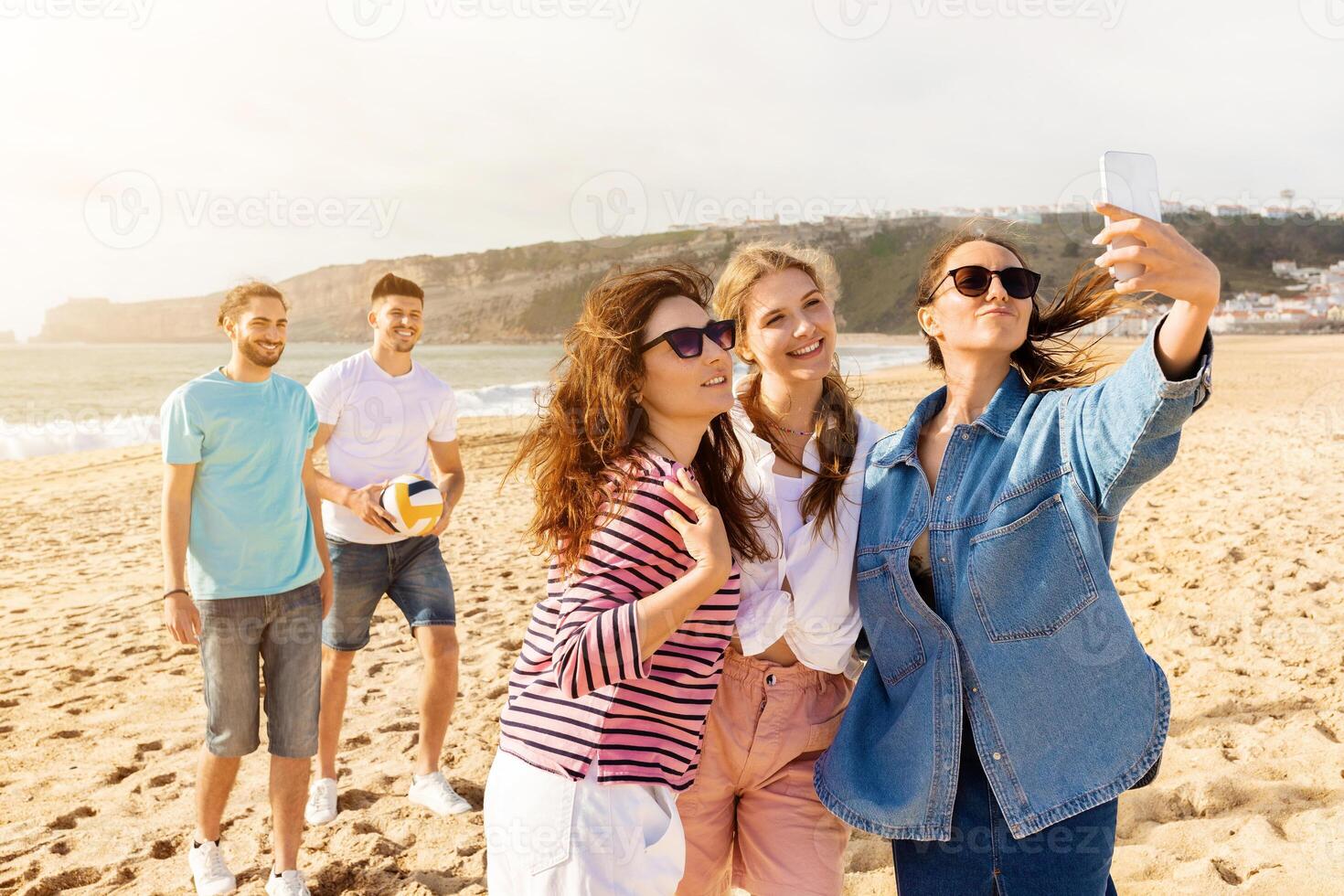 Four friends are having a great time at the beach, taking selfies while two others play volleyball in the background. The sun is shining beautifully over the sandy shore. photo