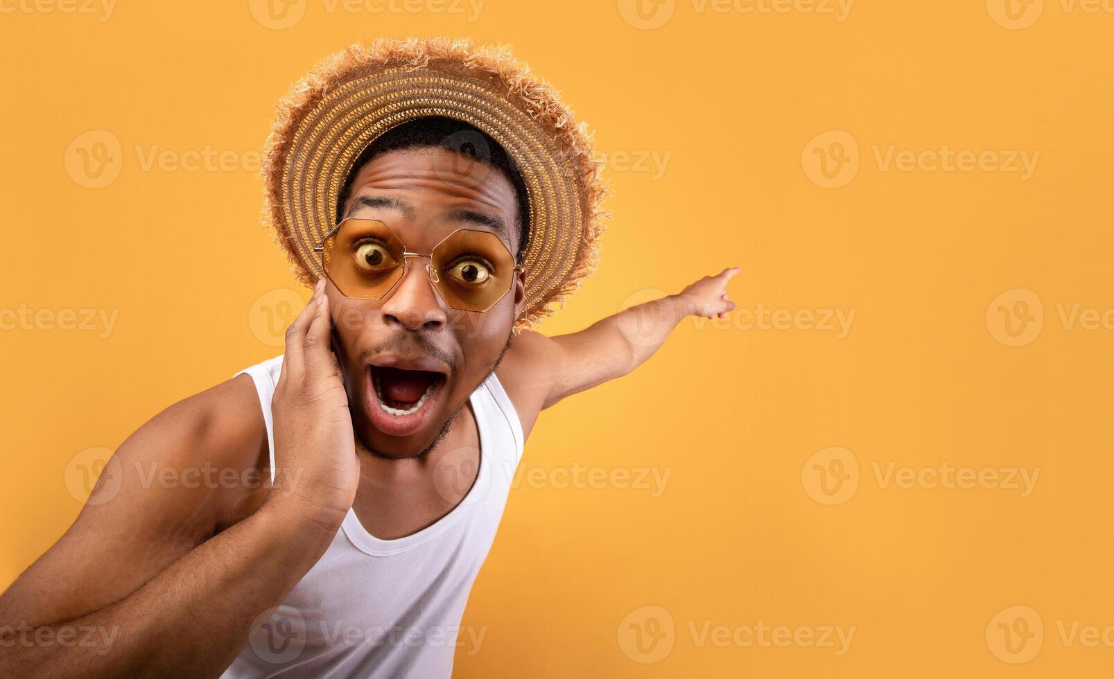 Summer sale. Shocked black guy in straw hat pointing at empty space on yelow studio background. Surprised African American man advertising your summertime product or service photo