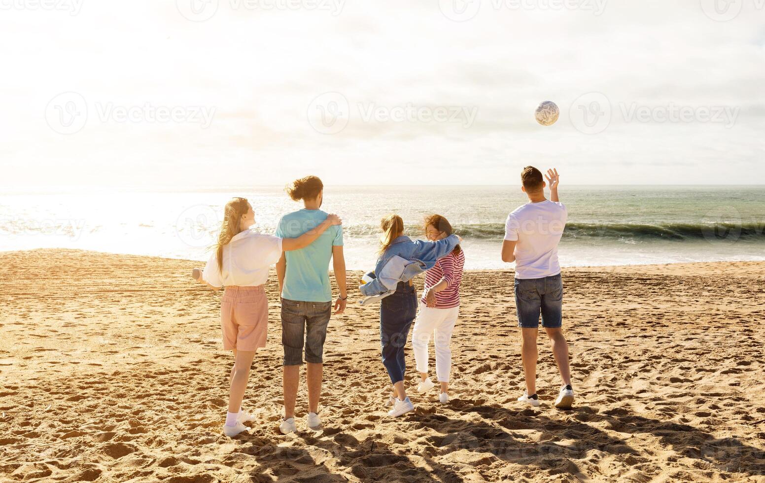A group of five friends stands on the sandy beach, enjoying a sunny day. They are playing with a ball and laughing as the waves crash gently behind them. photo