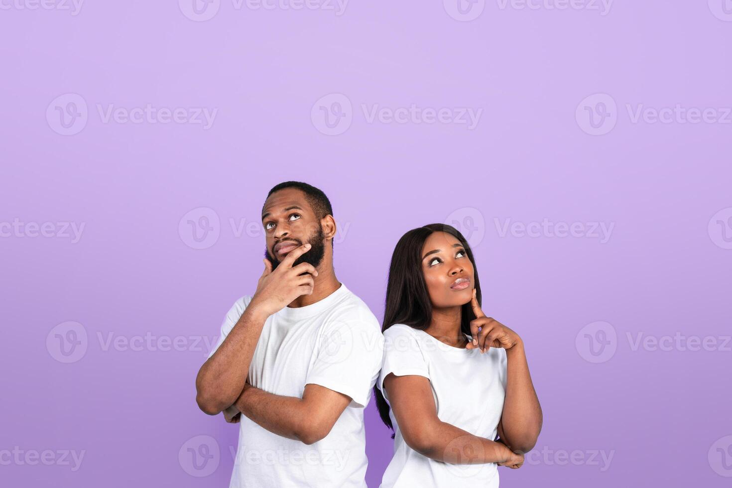 Portrait of thoughtful black guy and lady thinking about question and looking up, touching chin, planning future vacation and making decision, standing back to back on blue color studio wall photo