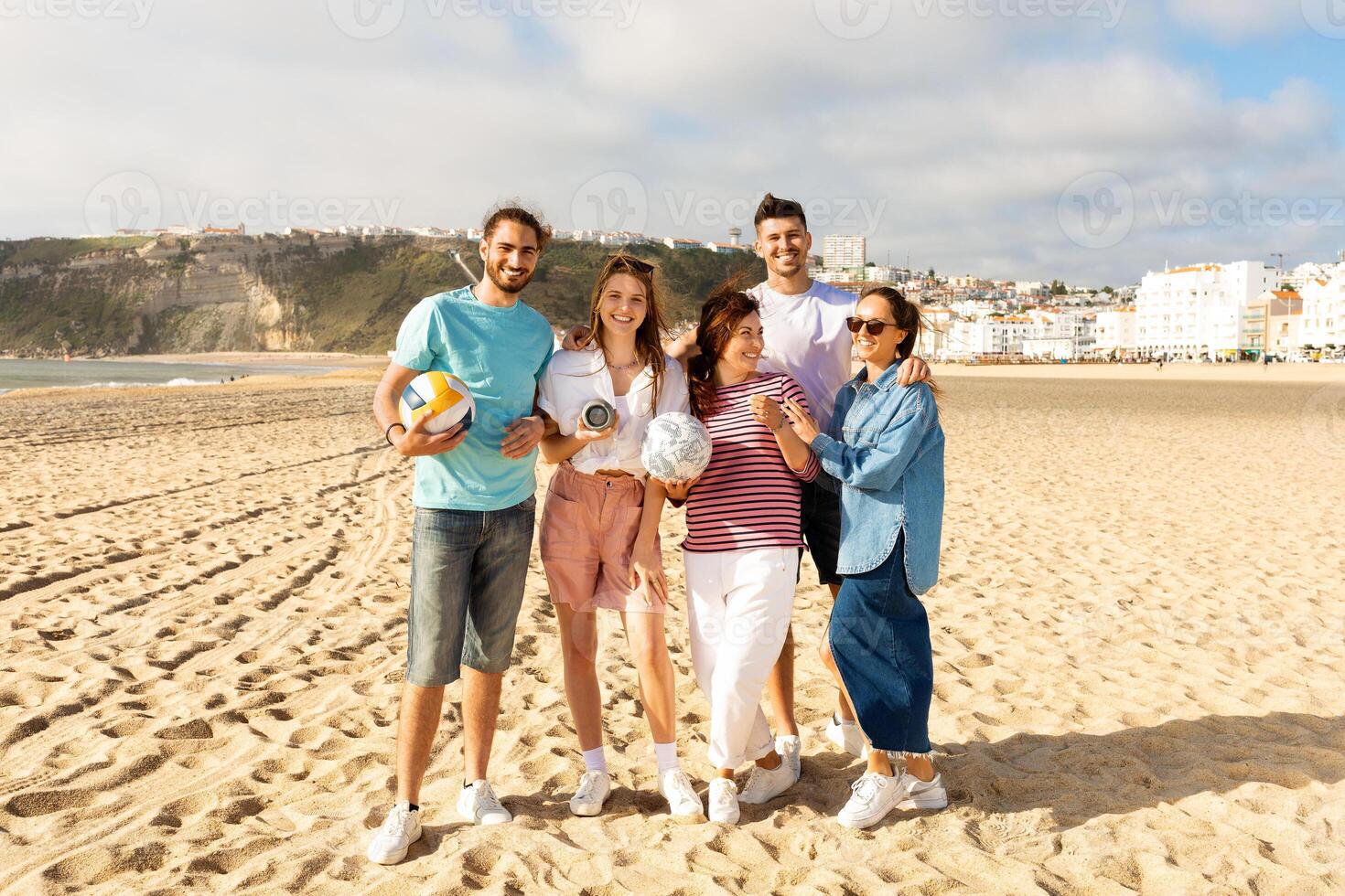 A cheerful group of five friends stands on the sandy beach, each holding sports equipment. They smile together in the warm sunlight, creating joyful memories by the sea. photo