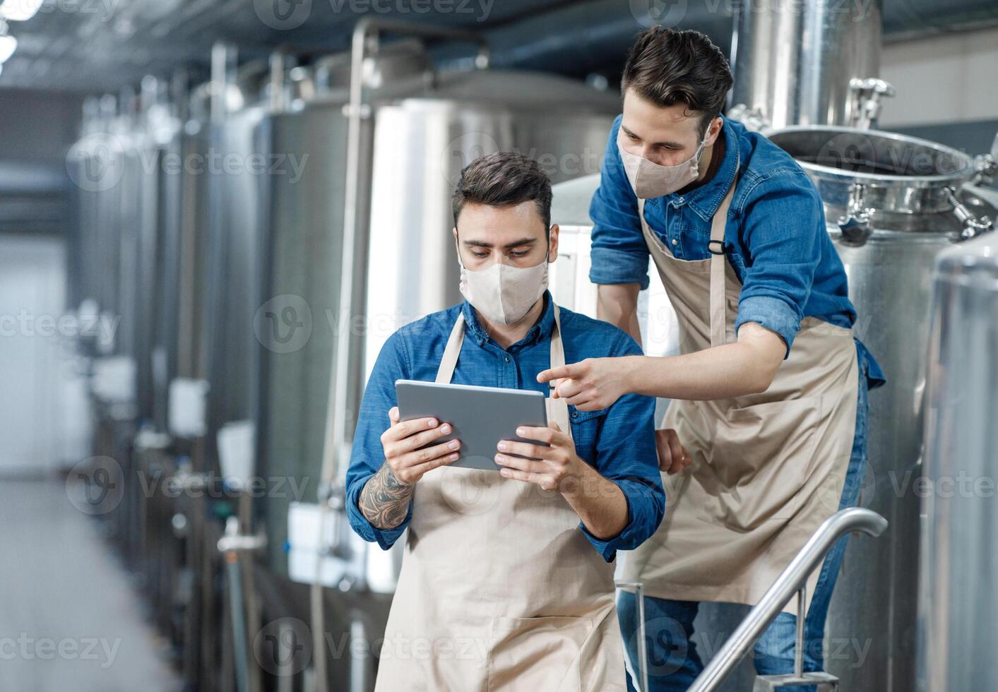 Brewery workers enter data at tablet and operate equipment. Young guys in protective masks and aprons discuss and work with craft beer tanks in interior of factory during COVID-19 epidemic, free space photo
