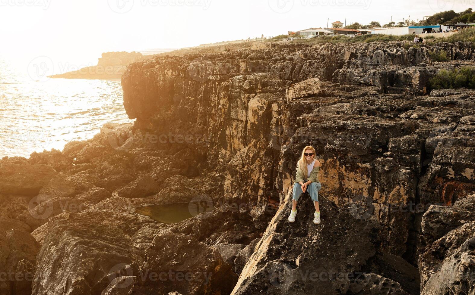 A woman relaxes on a rocky cliff, gazing towards the ocean as the sun sets behind her. The serene landscape showcases cliffs and a coastal town in the distance. photo