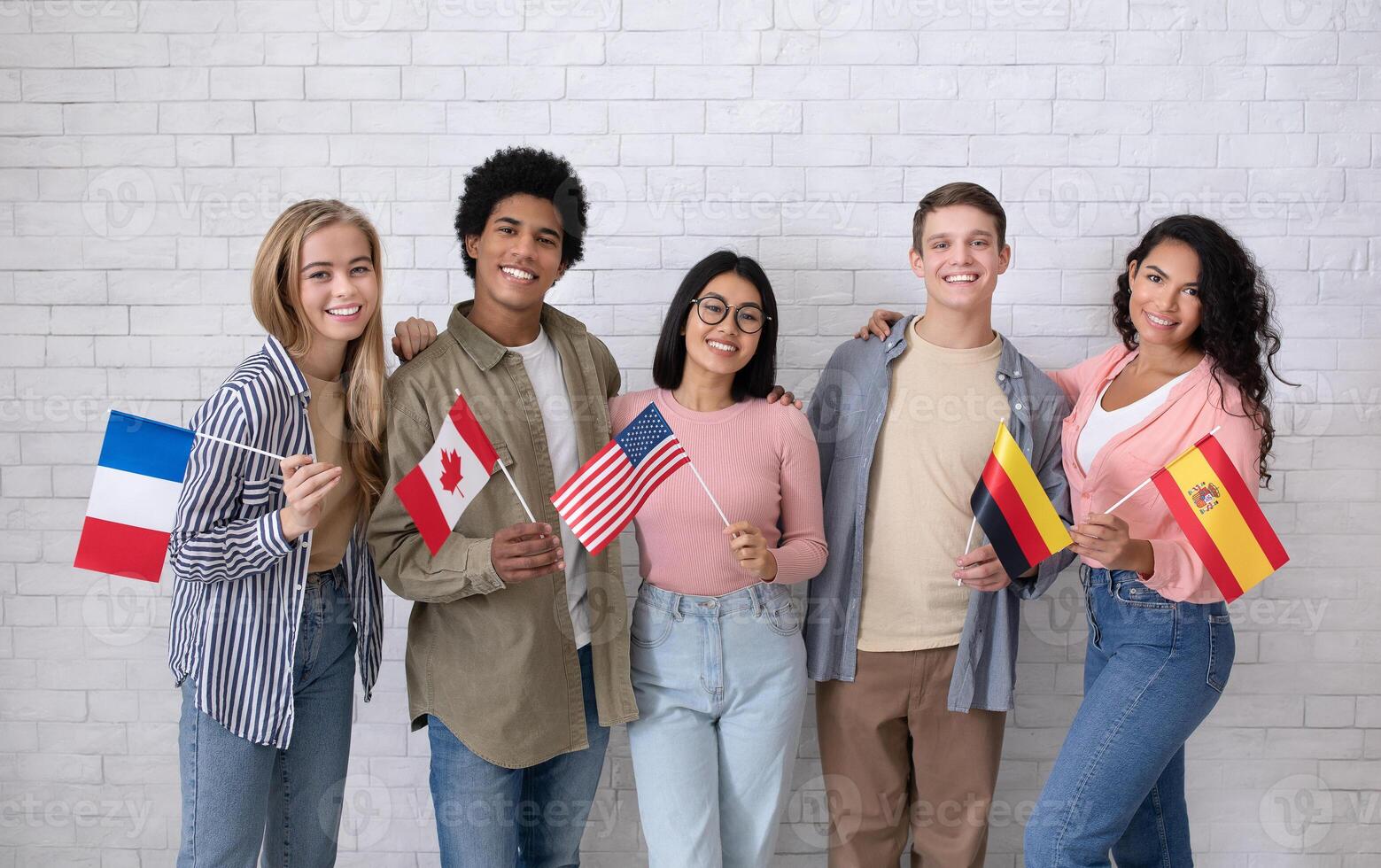 Immigrants of different countries and exchange students in language school. Smiling multiracial young people hold flags of France, Canada, USA, Germany and Spanish in classroom, studio shot photo