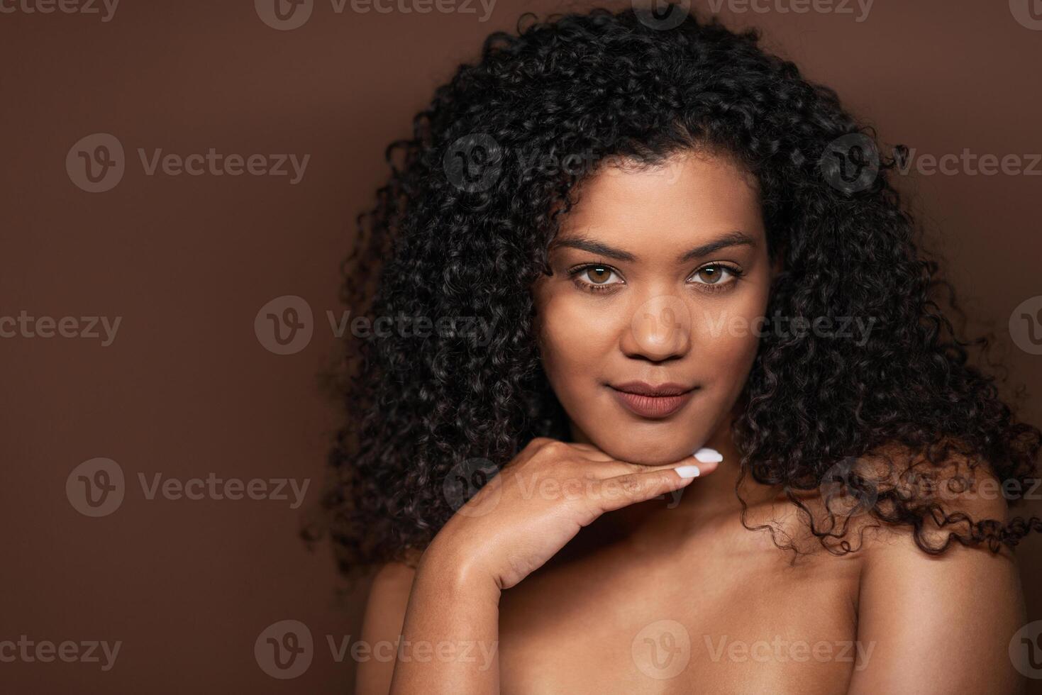 A woman showcases her natural beauty with striking curly hair while gently resting her chin on her hand. The warm brown backdrop highlights her features and confident expression. photo