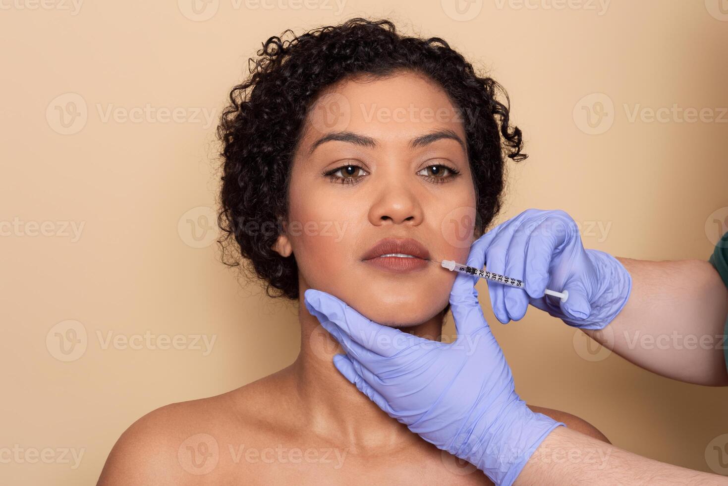 A woman with curly hair sits calmly while a practitioner administers a dermal filler to enhance her lips in a beauty clinic. The bright environment adds to the soothing atmosphere. photo