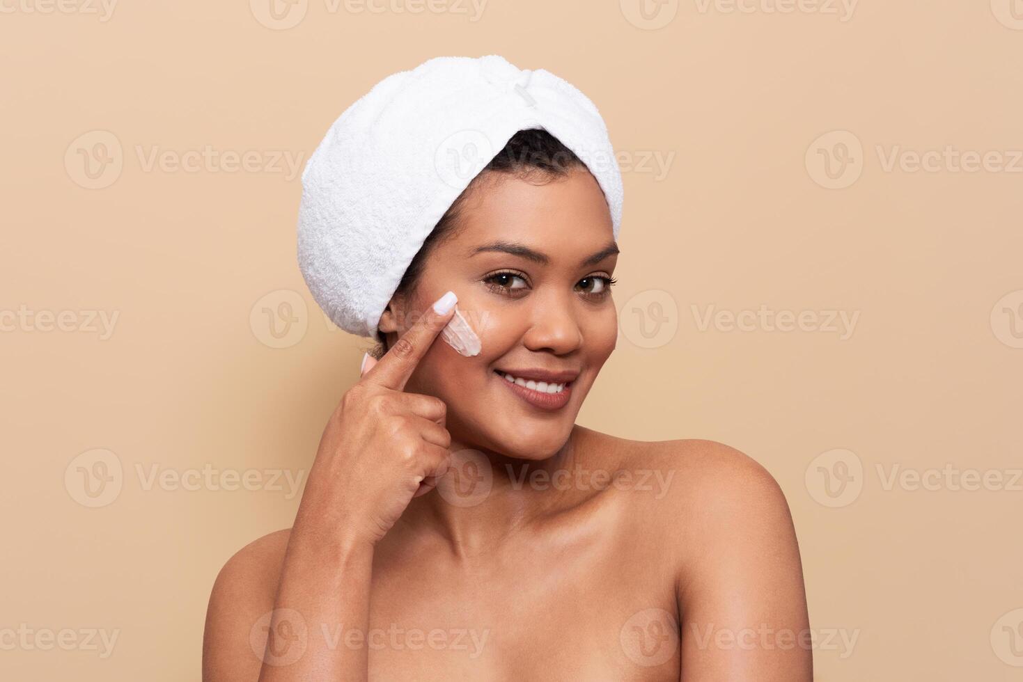 A young woman with a towel wrapped around her head is smiling while applying cream to her face. She is in a serene indoor setting, suggesting a skincare routine or self-care moment. photo