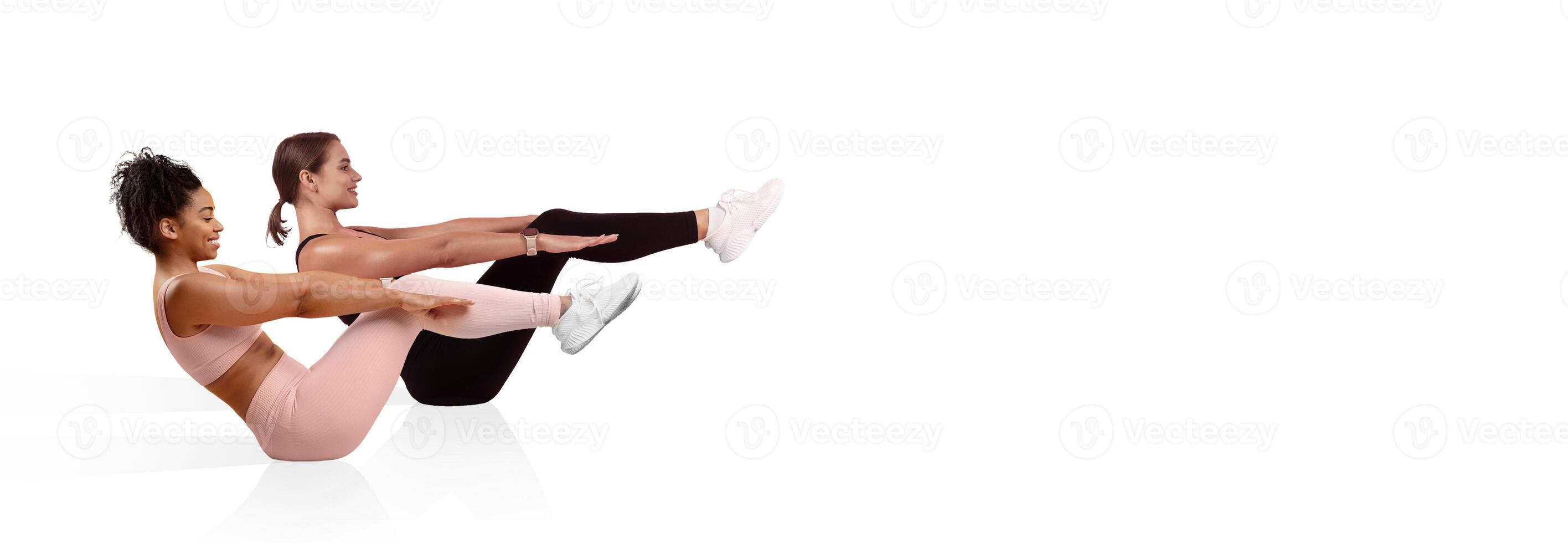Two women are engaged in synchronized core exercises, sitting with legs raised and leaning back. The brightly lit gym offers a clean and modern atmosphere for workout routines. photo