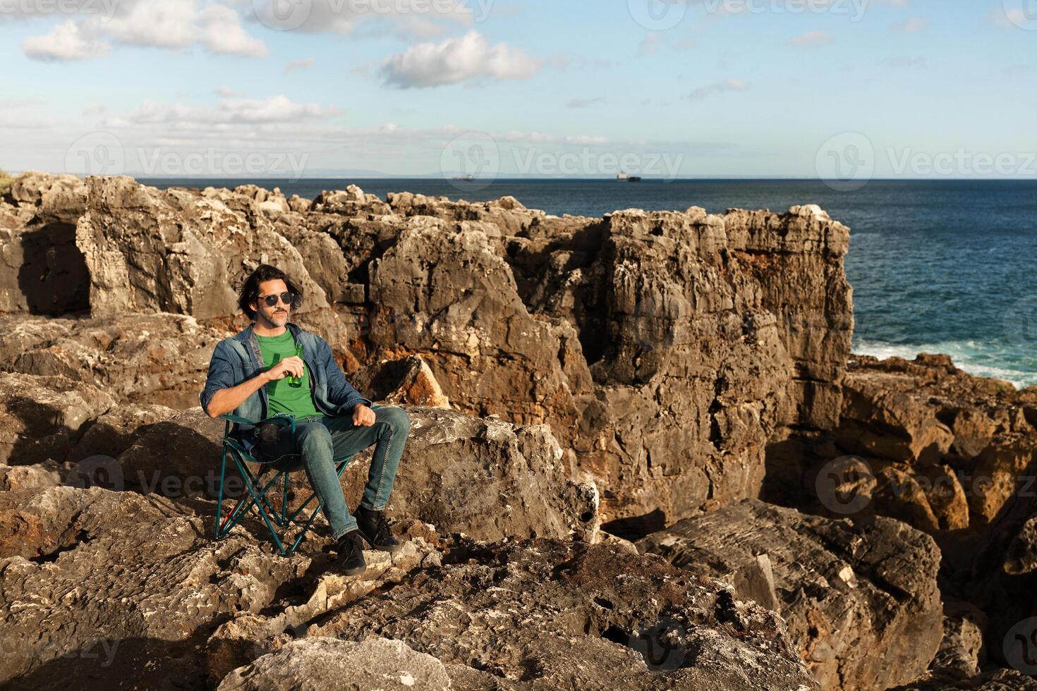 A man sits comfortably on a chair among rocky formations by the ocean, savoring a drink. He enjoys the serene coastal view under a clear sky during daytime. photo