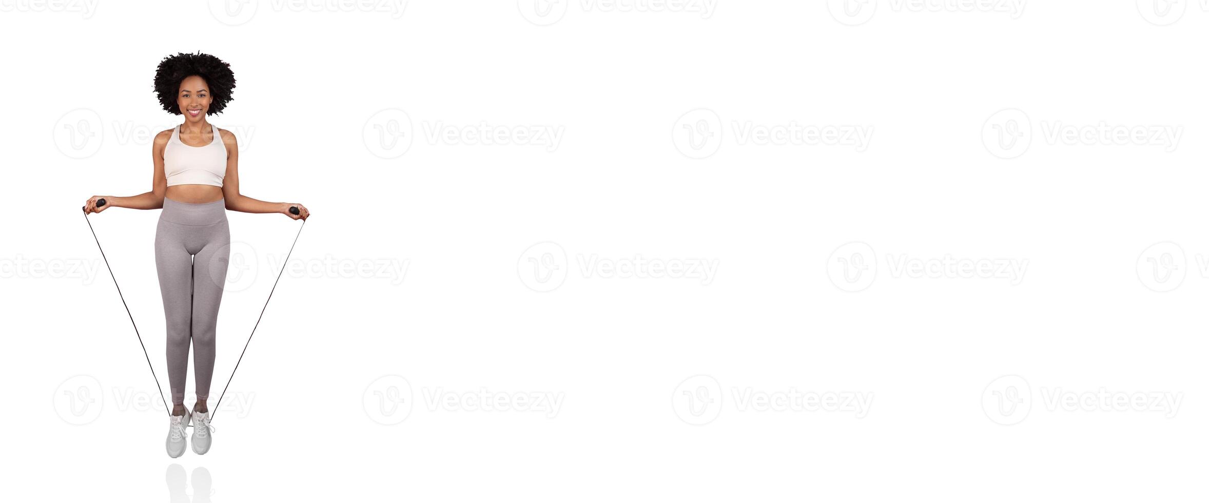 A woman is joyfully jumping rope in a bright indoor space. She wears a matching light outfit and shows a vibrant smile, embodying energy and wellness during her workout routine. photo