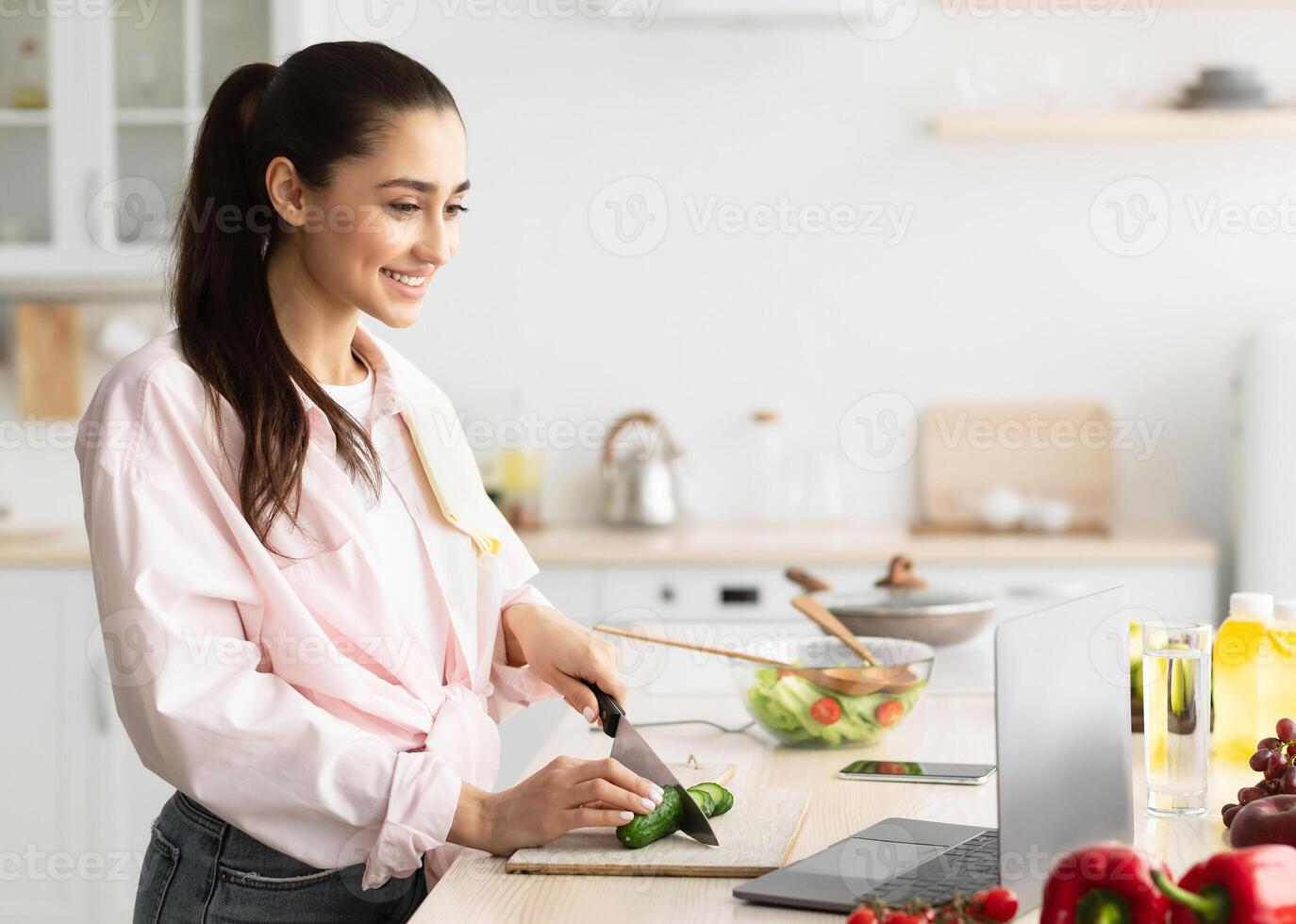 Cooking Courses Online. Portrait of positive cheerful woman watching blog on laptop, standing at kitchen and preparing healthy meal. Happy young housewife slicing cucumber following new recipe photo