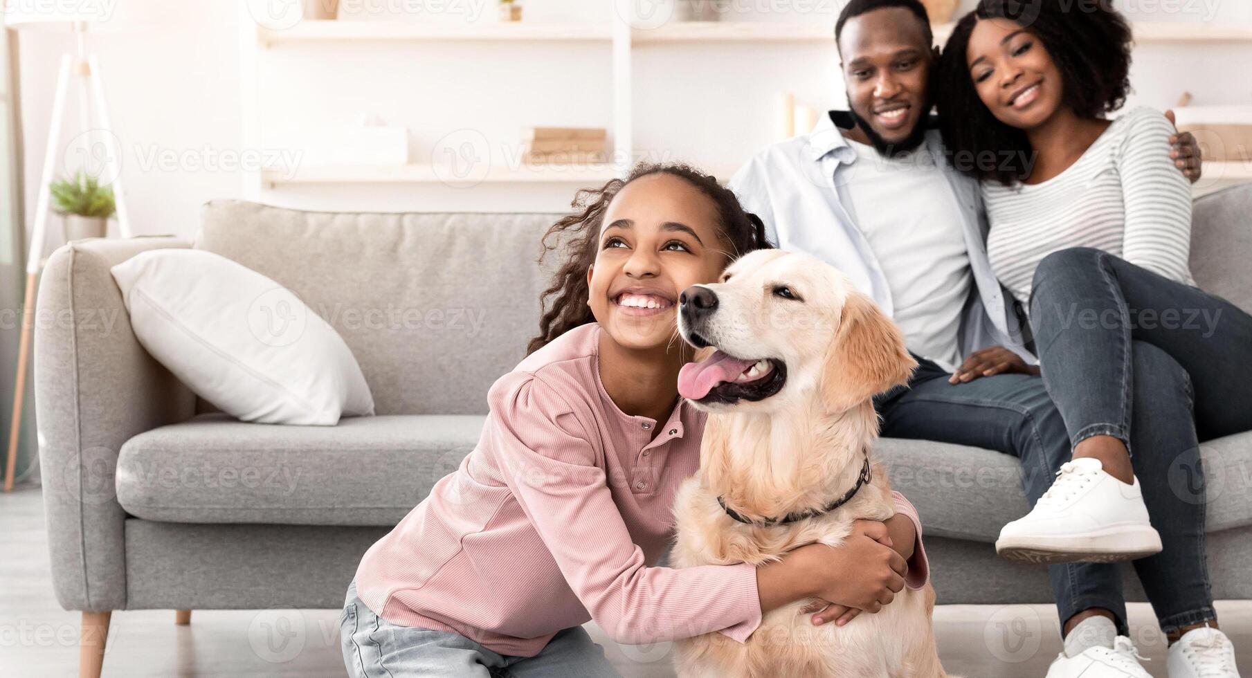 Man's Best Friend. Portrait of smiling black girl hugging her happy dog in living room at home, looking away, cheerful parents sitting on the couch and looking at daughter playing with pet photo