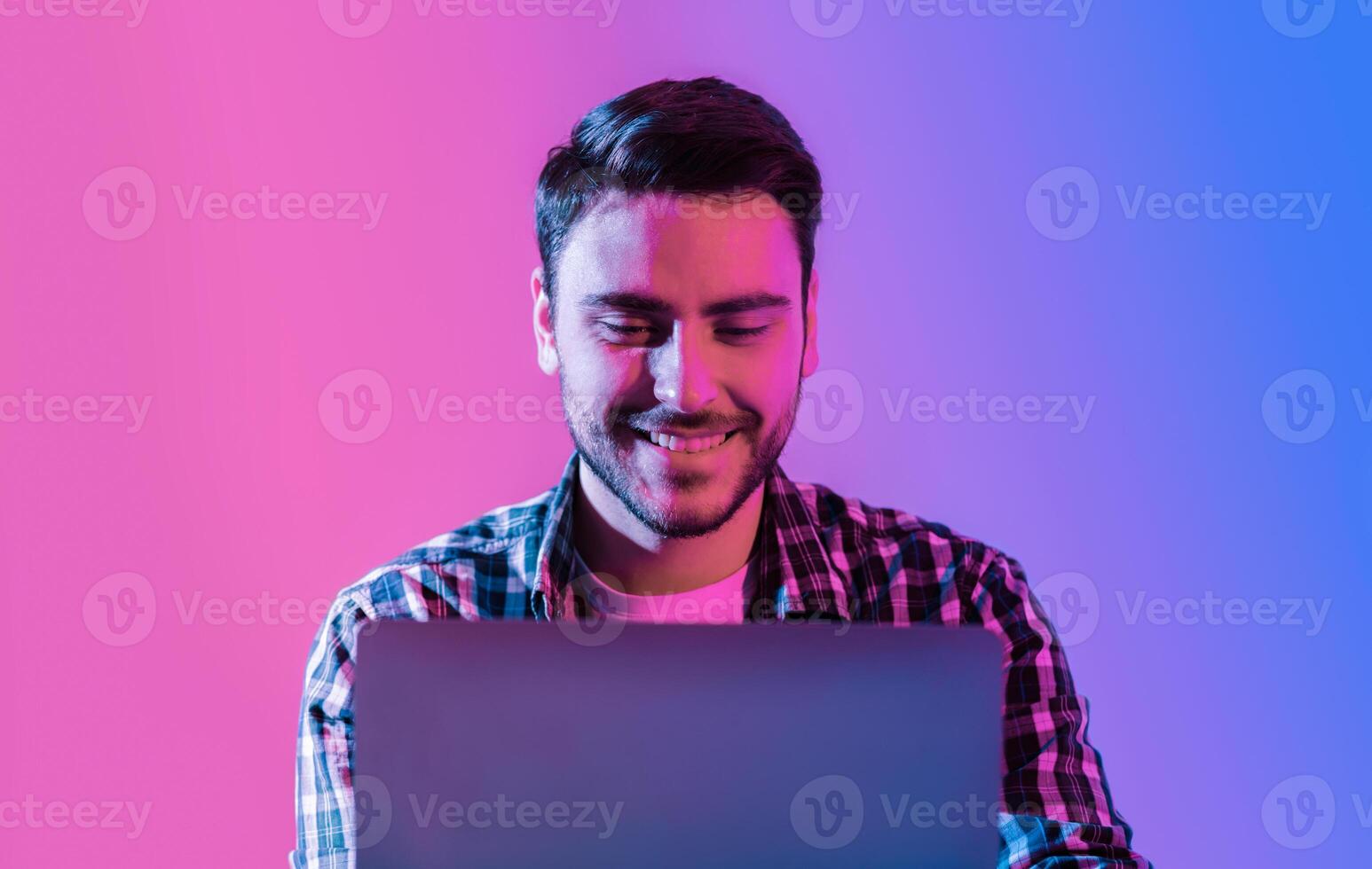 Freelancer at work. Young smiling man with laptop in neon studio shot photo
