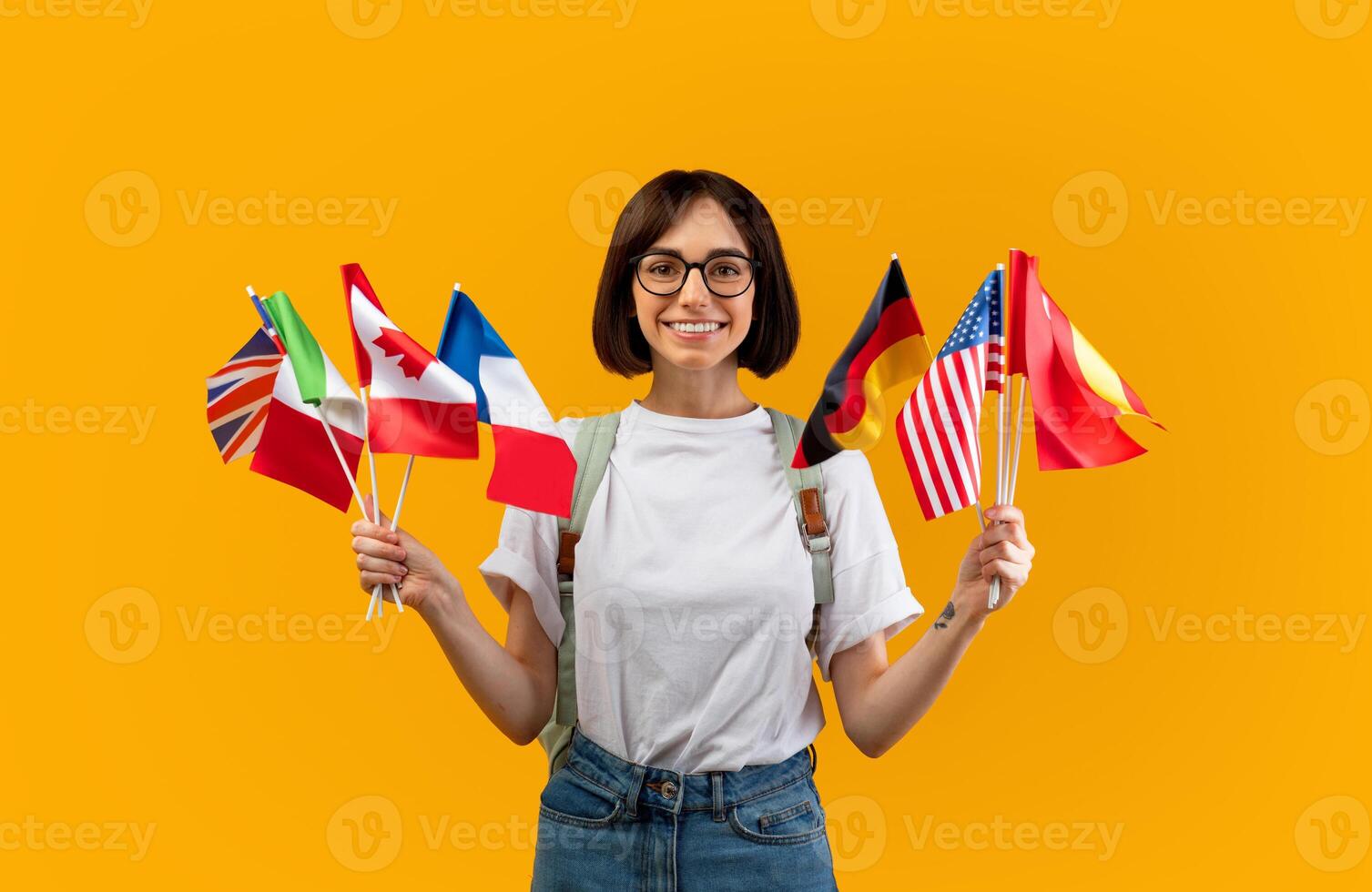 Happy lady showing bunch of diverse flags cheerfully smiling at camera over yellow studio background, excited female student recommending foreign language studying school photo