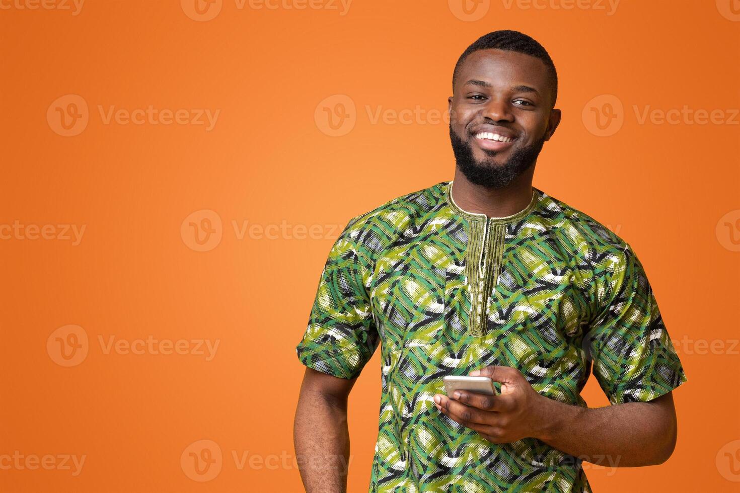 Smiling black guy in authentic african costume using mobile phone, yellow background, copy space photo
