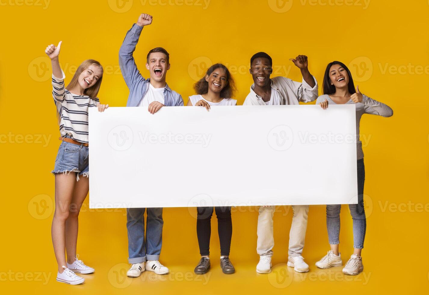 Happy smiling group of multiracial students standing together and displaying white blank placard, yellow studio background photo