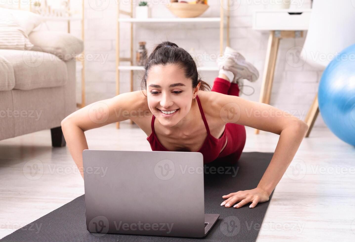 Young athletic woman exercising at home, doing push ups in front of laptop, watching online workout tutorial photo