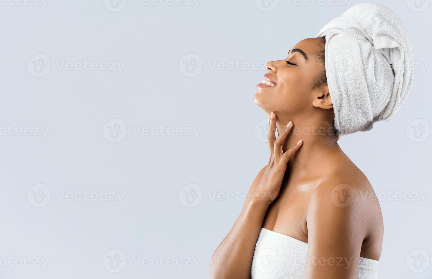 Side view of joyful young african woman wrapped in white bath towels after shower, enjoying her silky skin, empty space, grey studio background, discounts at spa or sauna photo