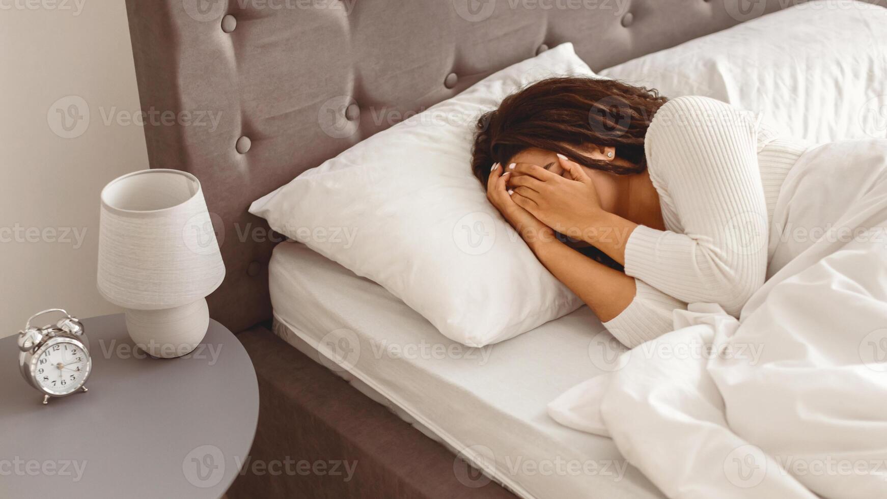 Depression Concept. Portrait of upset African American woman crying, covering face with hands, lying in bed under blanket. Sad black female feeling lonely, banner. Alarm on the table photo