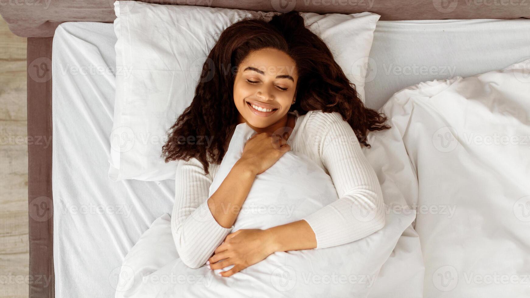 Good Morning. Above top high angle view of smiling young black woman waking up, holding and hugging pillow, feeling good after sleeping well, charming curly lady lying on the white bedsheets at home photo