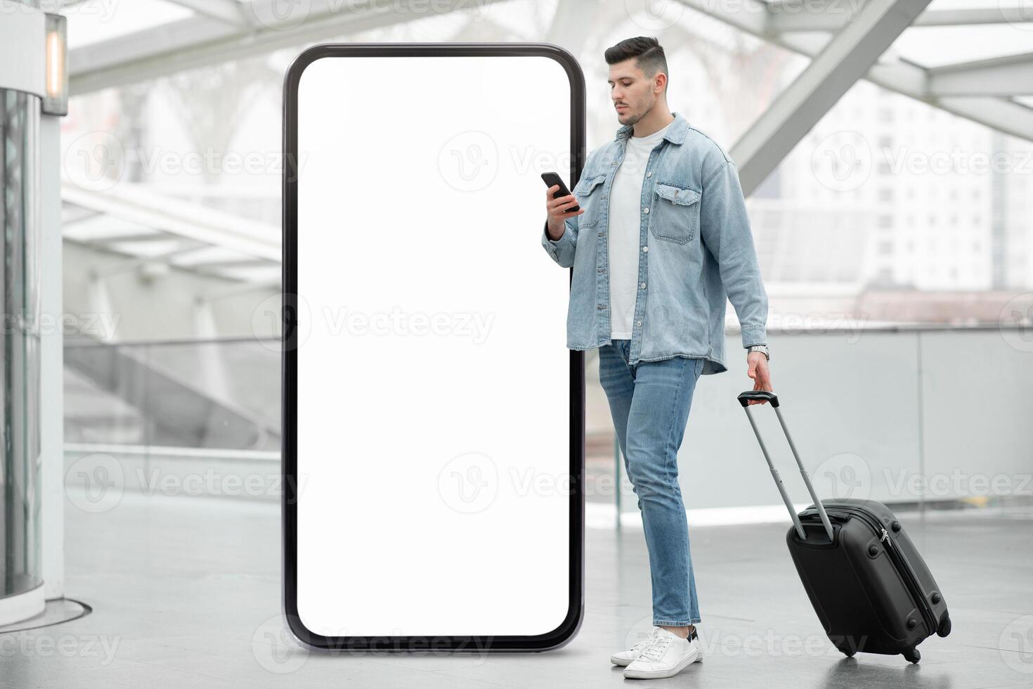 A traveler waits at a modern transit hub, checking a smartphone while standing beside a large blank billboard. The area features sleek architecture and a contemporary design. photo