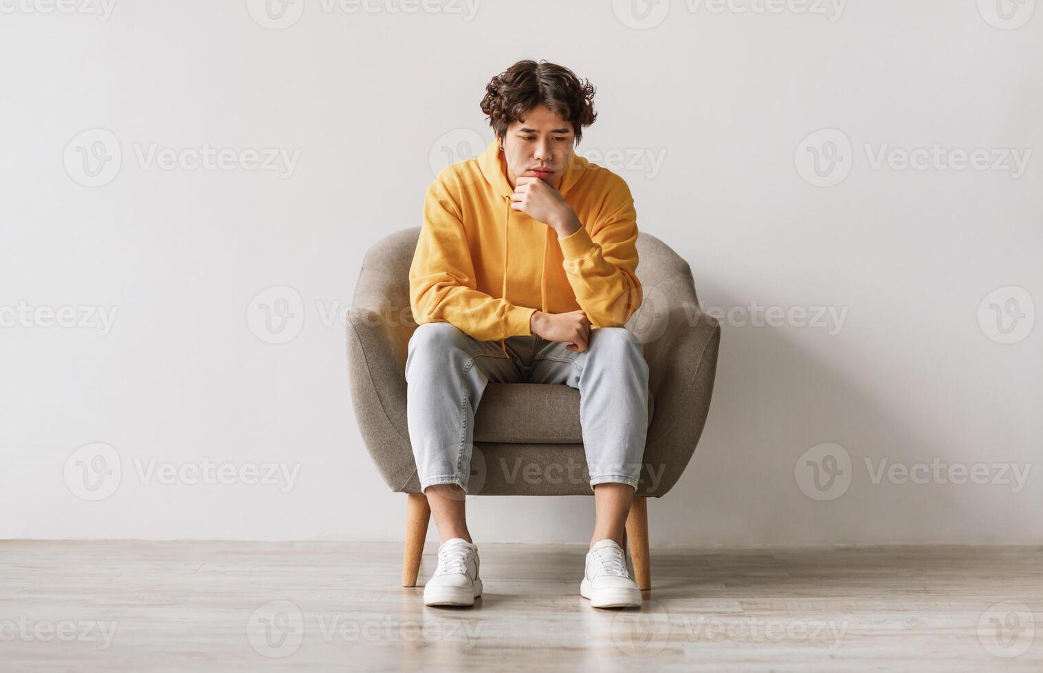 Negative emotions. Young Asian man sitting in armchair with thoughtful face expression, feeling sad or tired, having problem, suffering from depression or stress against white studio wall photo