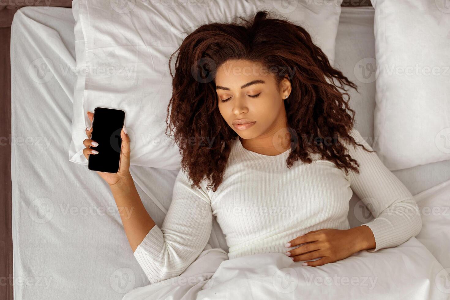 Above Top High Angle View Of African American Woman Sleeping Holding Smartphone Resting In Bed At Home. Always In Touch, Busy And Stressed Modern Life, Mobile Phone And Gadget Addiction Concept. photo