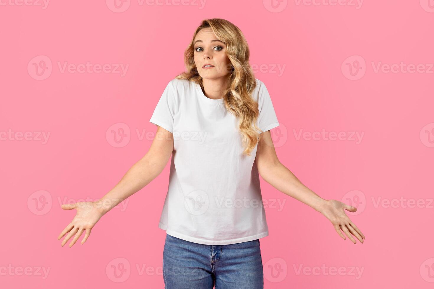 A young woman stands with her arms raised and a puzzled expression on her face. The background is a bright pink, creating a fun and lively atmosphere. She wears a plain white t-shirt and blue jeans. photo