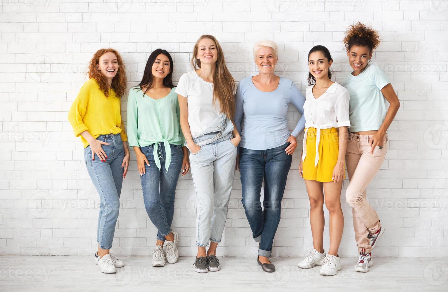 Diverse Ladies Of Different Age Posing Standing Over White Brick Wall Smiling To Camera. Full Length photo