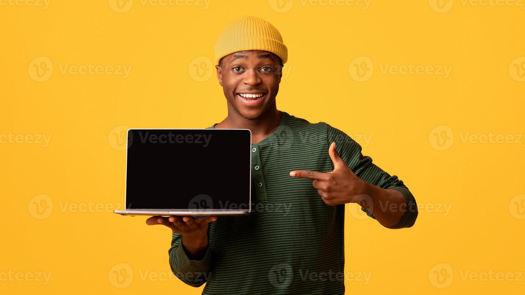 Cool Website. Cheerful African American Guy Pointing At Laptop Computer With Black Blank Screen, Demonstrating Free Copy Space For Your Design, Standing Isolated Over Yellow Background, Mockup photo
